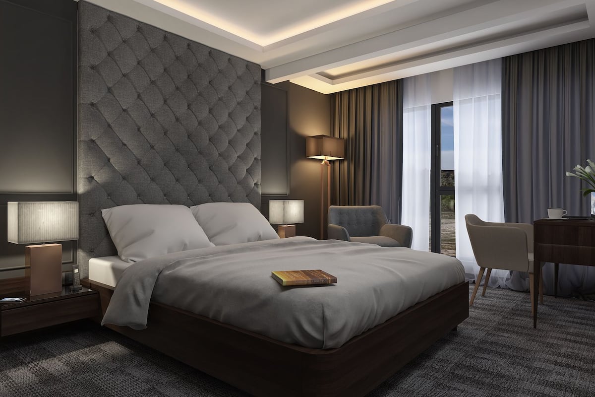 A modern hotel room with a large bed, two bedside tables with lamps, a desk with a chair, and a view of the outdoors.