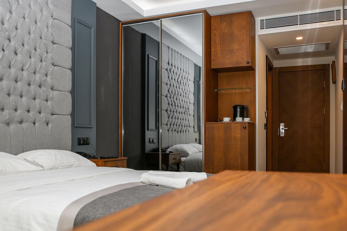 A modern hotel room with a tufted headboard, a large mirror, a wooden wardrobe, and a bedside table with a coffee maker.