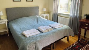 A neatly made bed with a quilted blue bedspread and folded white towels, a bedside table with a lamp, and two framed pictures on the wall. - Aspa Herrgård (Aspabruk)
