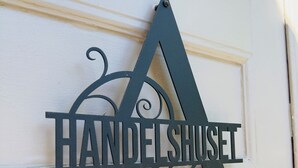 A metal sign with the letters "A" and "HANDELSHUSEL" mounted on a white wall. - Aspa Herrgård (Aspabruk)