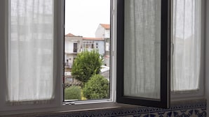 A window with white curtains, revealing a view of buildings and a tree outside. - Casa Velha Guesthouse (Porto)