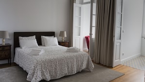 A neatly made bed with white floral bedding, two bedside lamps, and a view of an open door with curtains. - Casa Velha Guesthouse (Porto)