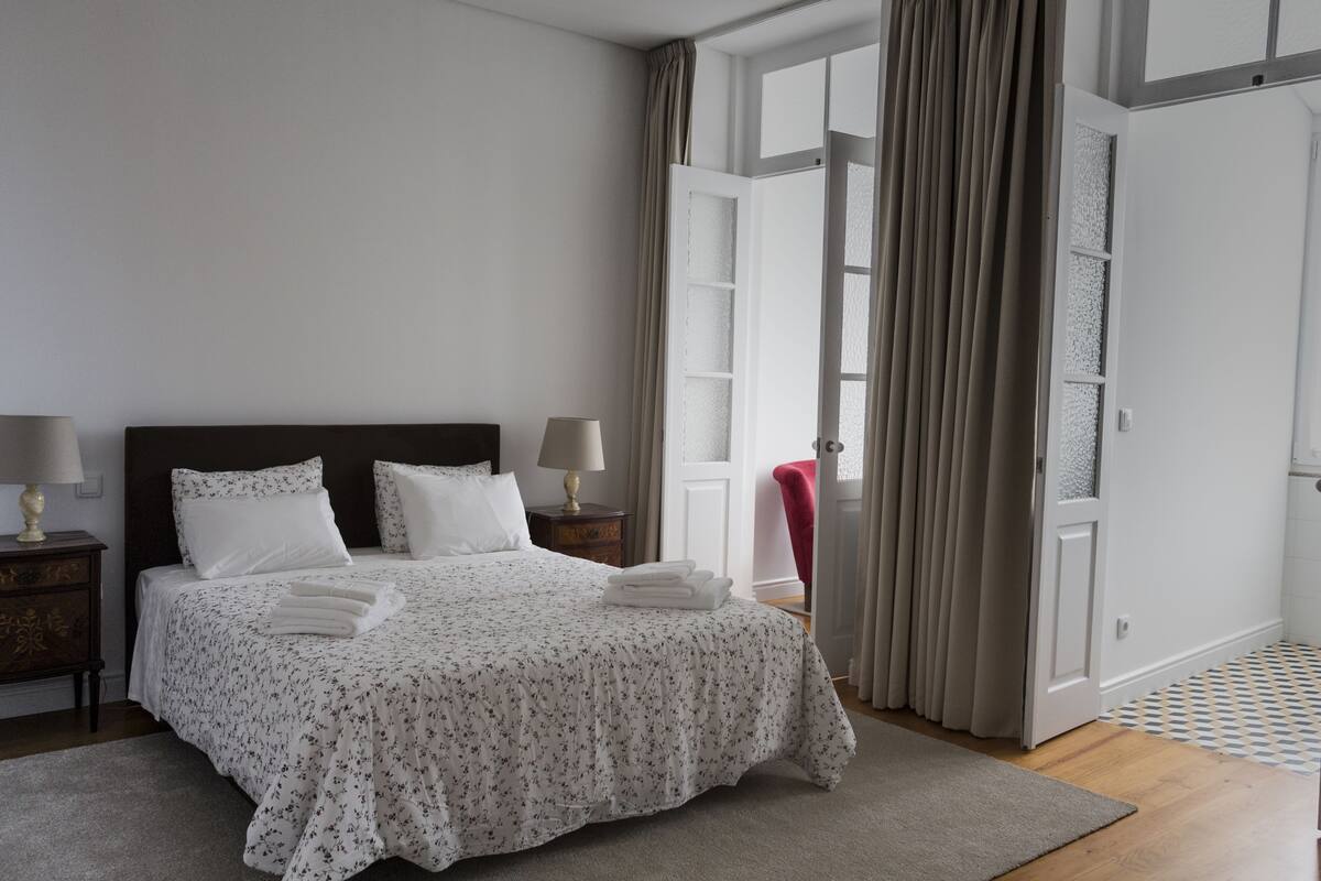 A neatly made bed with white floral bedding, two bedside lamps, and a view of an open door with curtains.