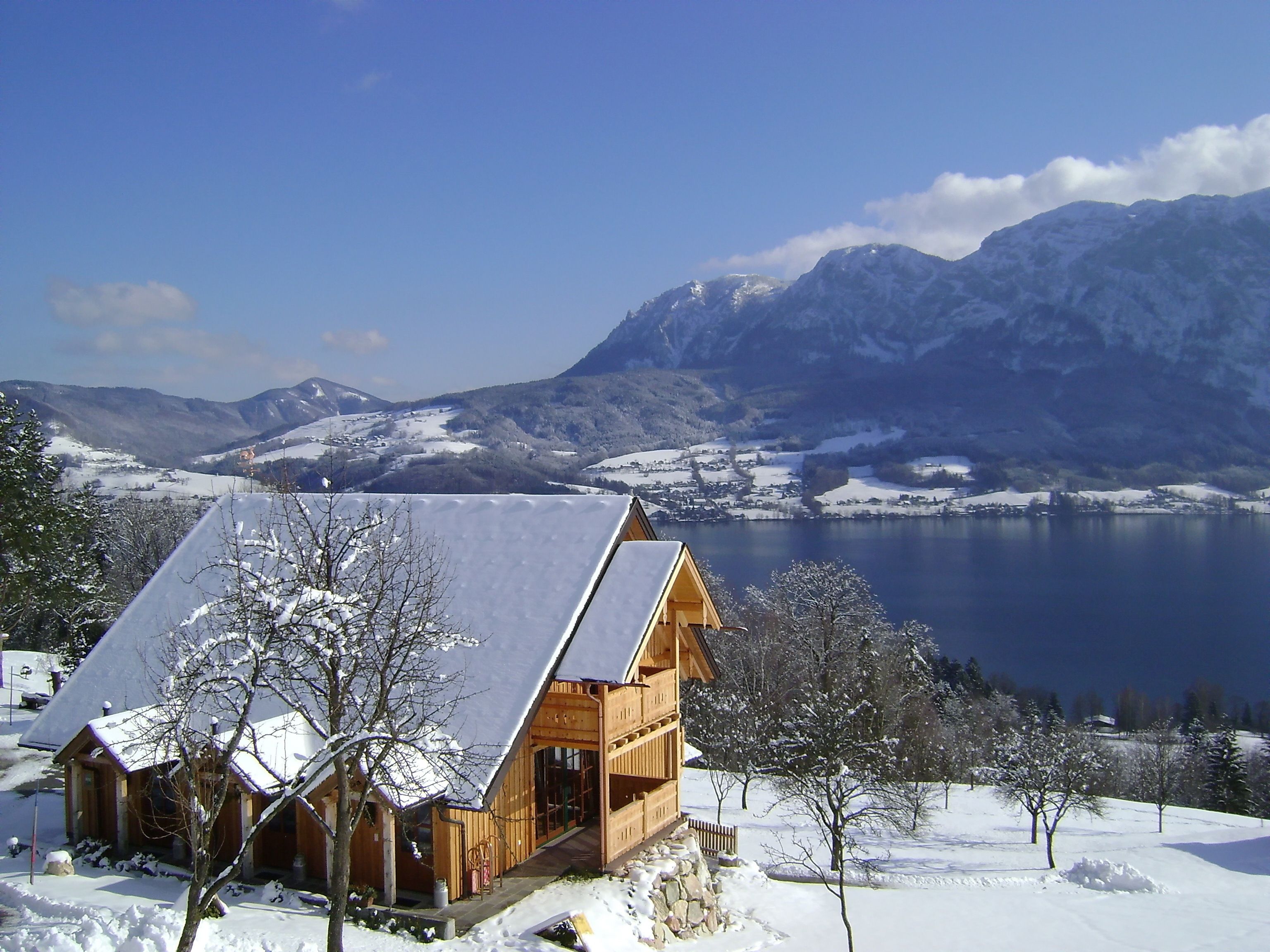Una cabaña de madera con techo inclinado, rodeada de árboles nevados y un lago, con montañas al fondo.