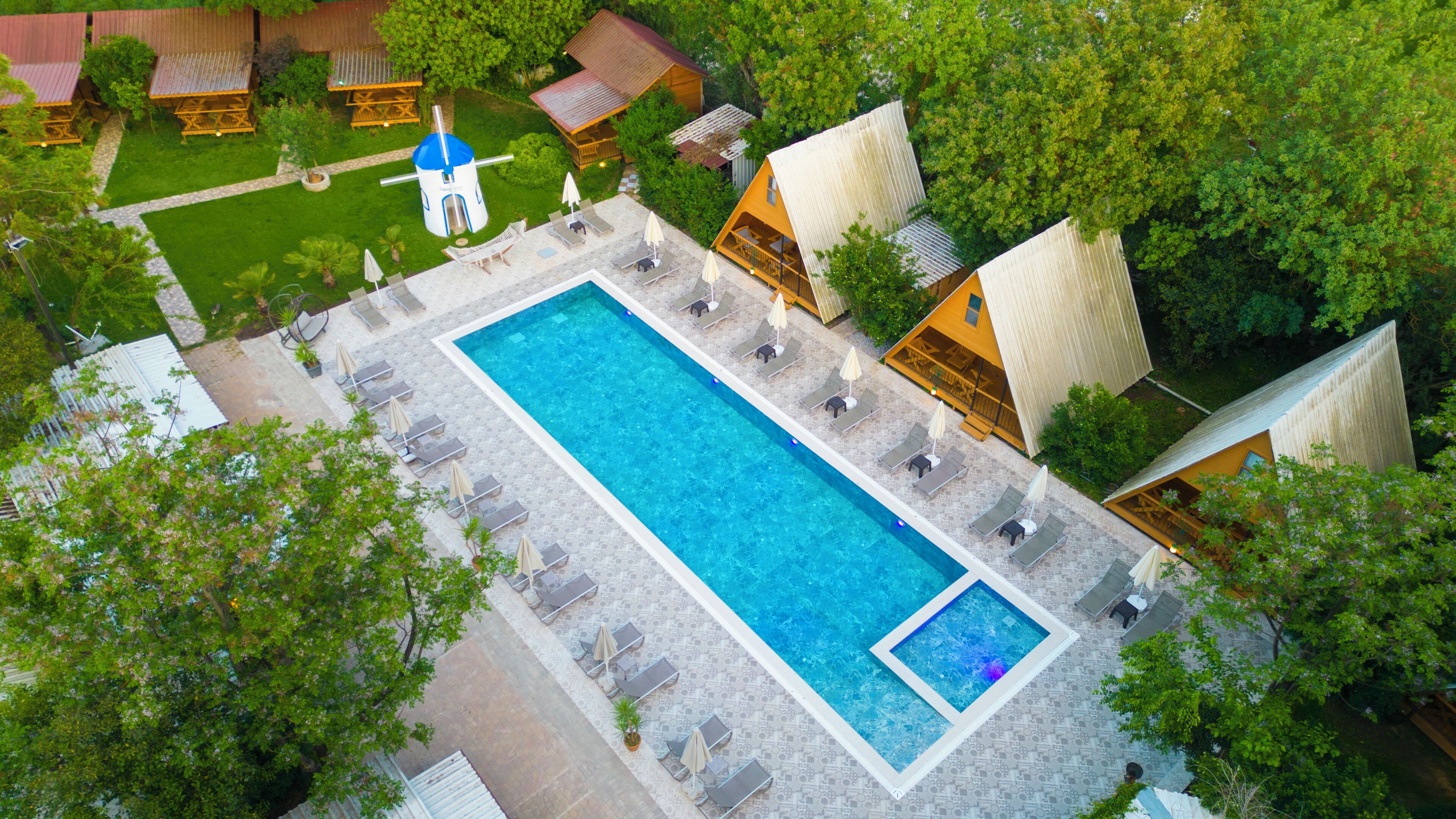 Vista aérea de un resort con una piscina rectangular rodeada de sillas de descanso y sombrillas, cabañas de madera y abundante vegetación.