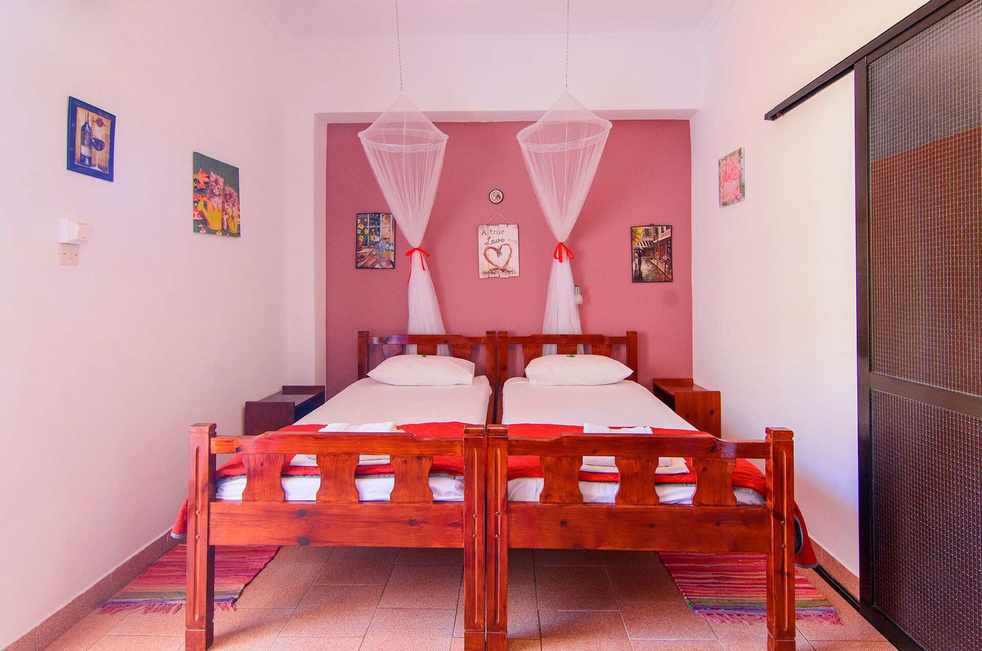Uma cama de casal com estrutura de madeira e roupa de cama branca, duas redes mosquiteiras e um tapete vermelho no chão.