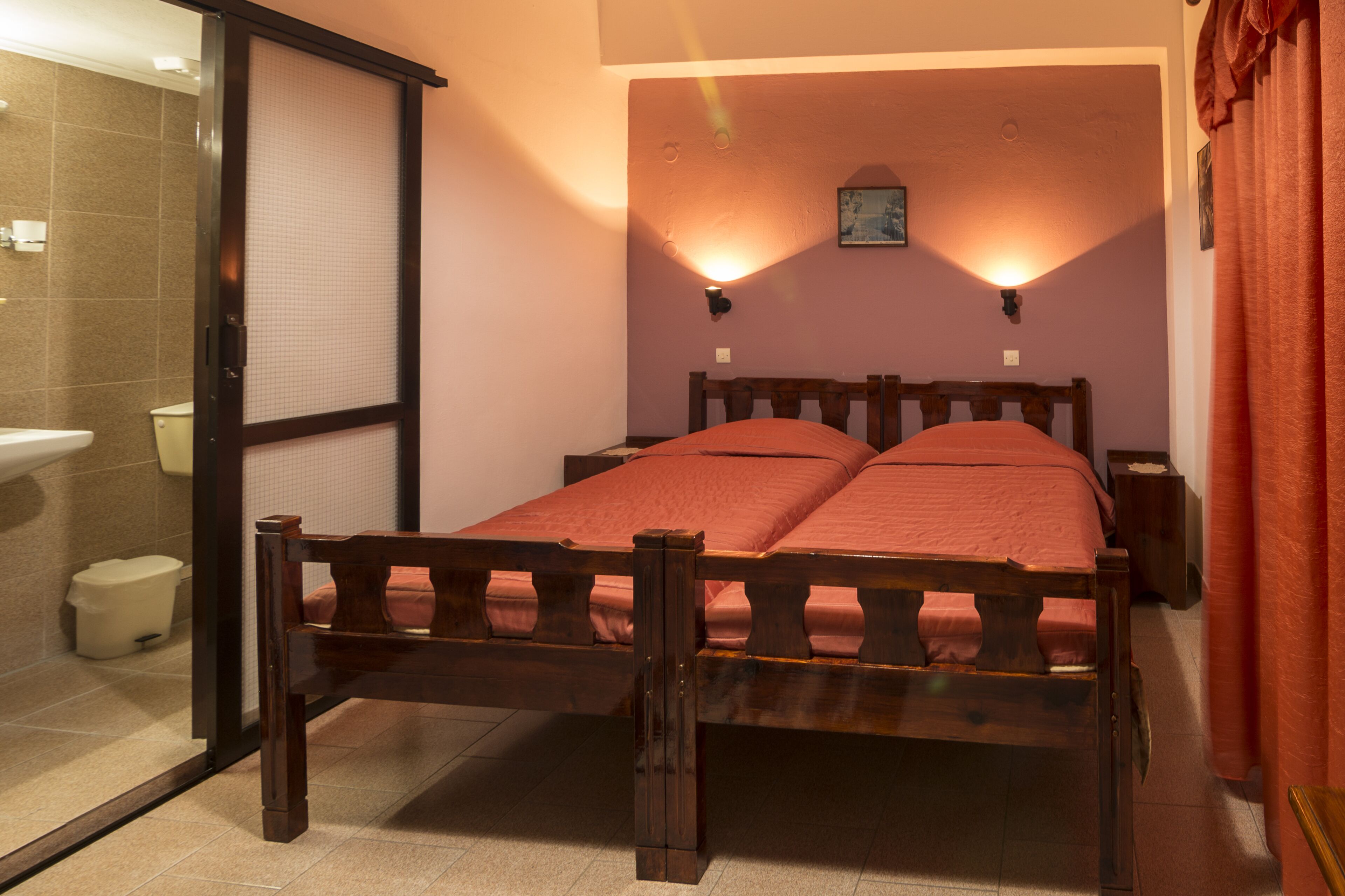 A bedroom with a wooden bed, two bedside tables, a nightstand, a wall-mounted light, and a bathroom visible through an open door.