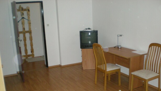 Una habitación pequeña y con pocos muebles, con un escritorio de madera, dos sillas, una cama y un televisor.