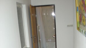 Un pasillo estrecho con piso de madera, una puerta que da a un baño con cortina de ducha y una ventana a la izquierda.