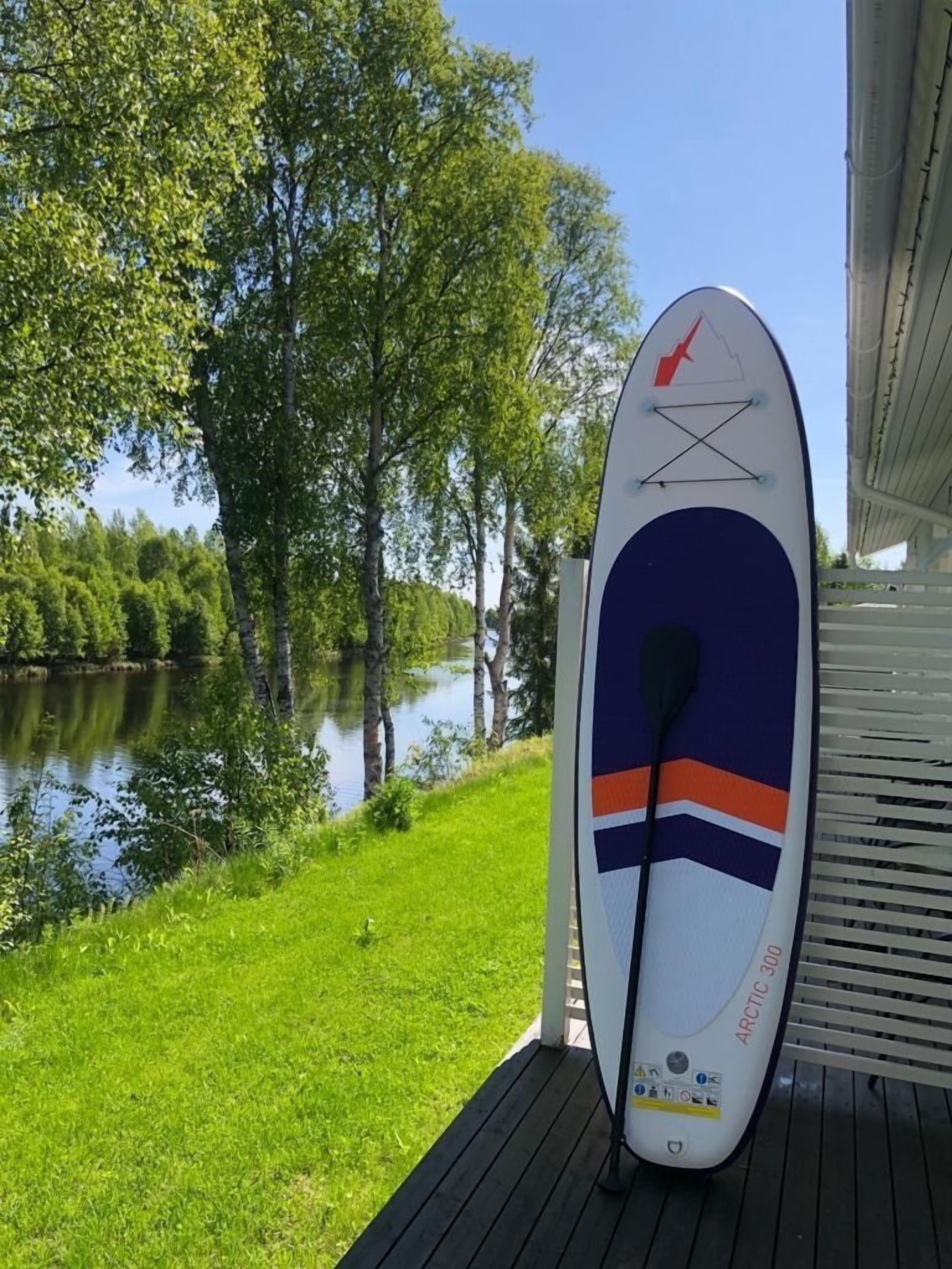 Une planche de surf sur une terrasse en bois, au bord d’un lac, avec des arbres en arrière-plan.