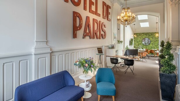 Un vestíbulo de hotel con un gran letrero que dice "HOTEL DE PARIS", un candelabro, zona de estar con sofás azules y un mostrador de recepción.