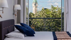Un dormitorio con cama, almohadas y vista a una torre del reloj a través de la barandilla de un balcón.