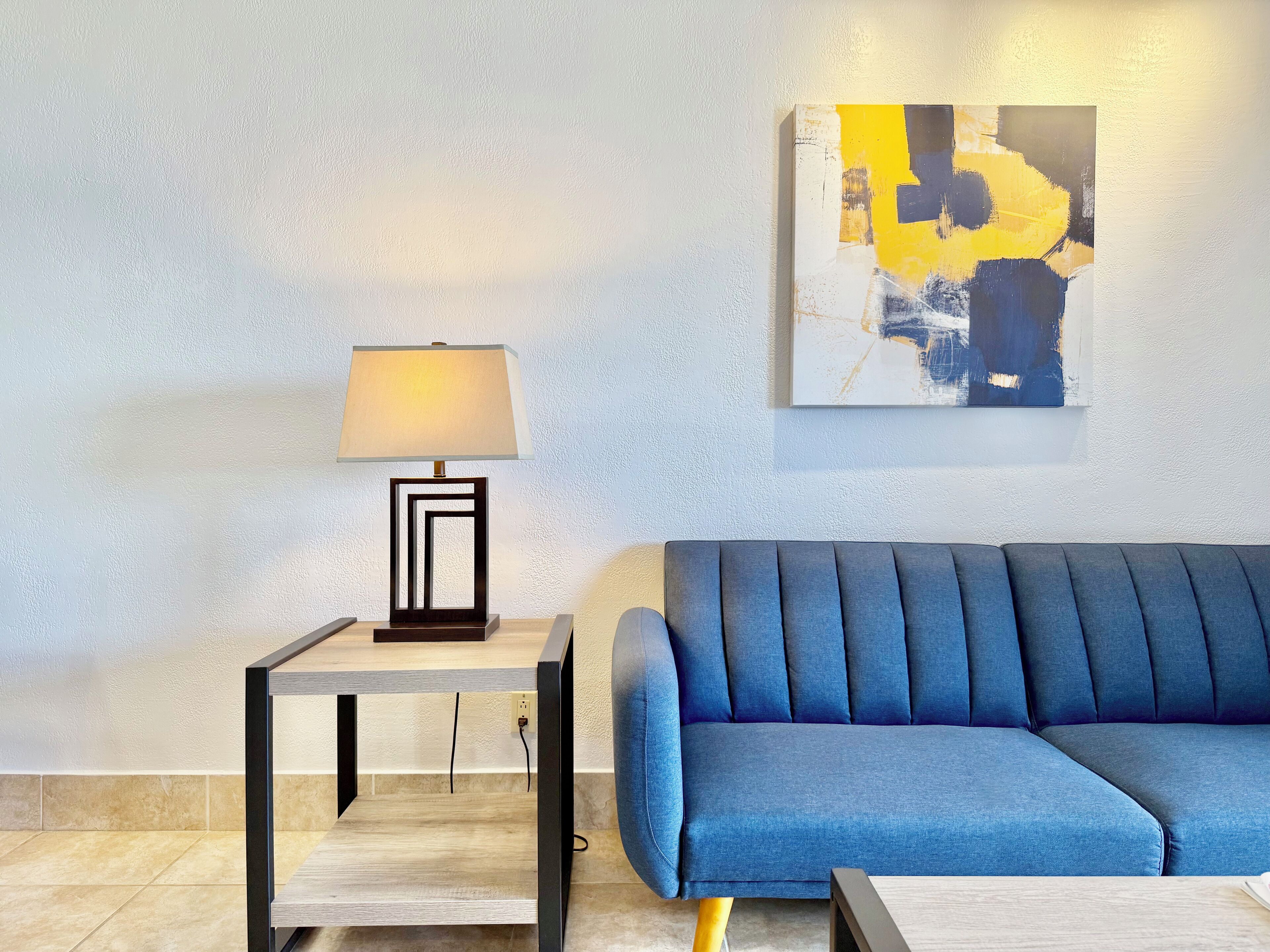 A modern living room with a blue sofa, a side table with a lamp, and a wall-mounted abstract painting.
