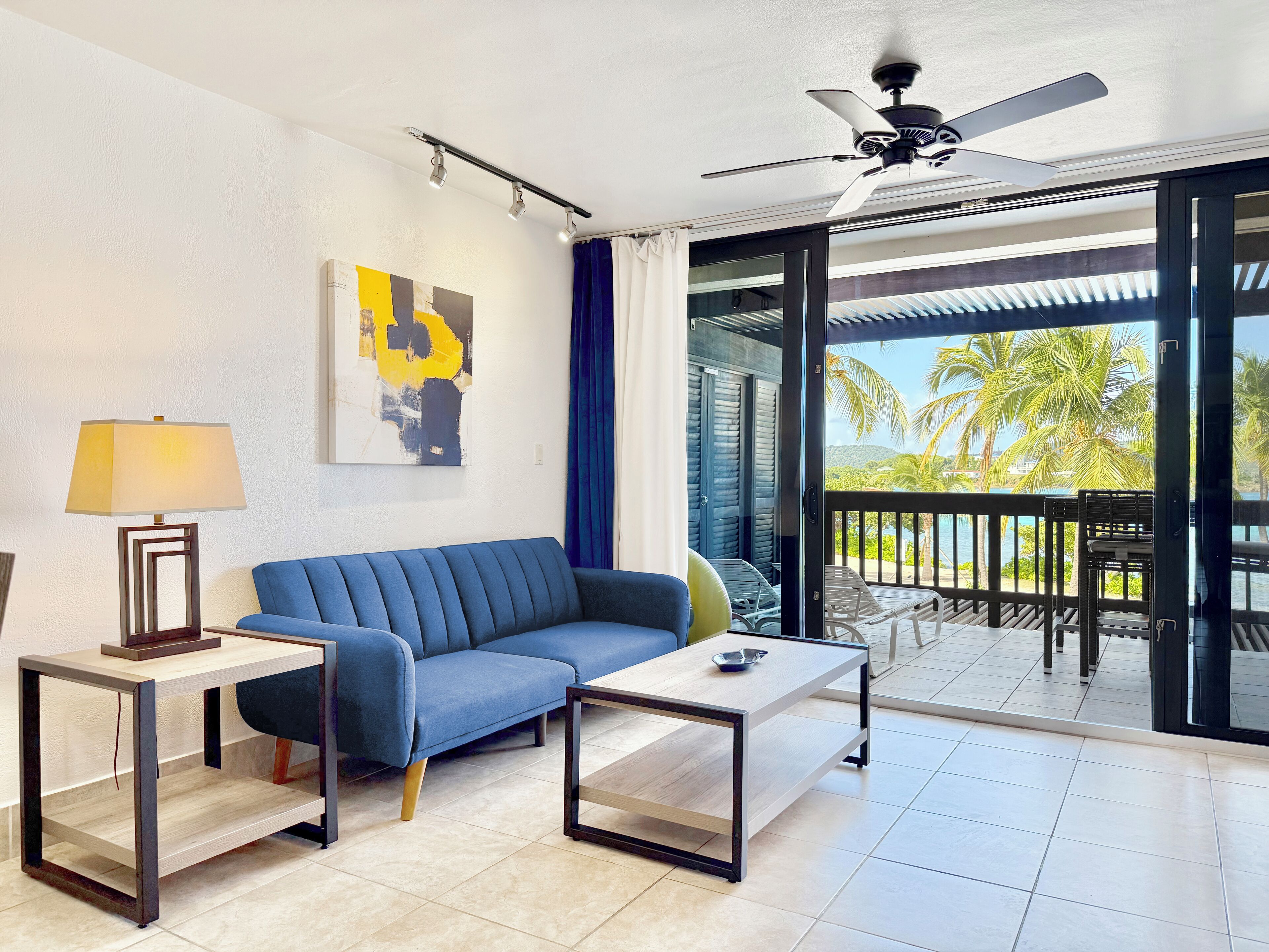 A modern living room with a blue sofa, a coffee table, and a floor lamp. There is a painting on the wall and a view of palm trees and water outside.