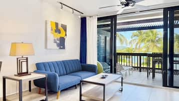 A modern living room with a blue sofa, a coffee table, and a floor lamp. There is a painting on the wall and a view of palm trees and water outside.