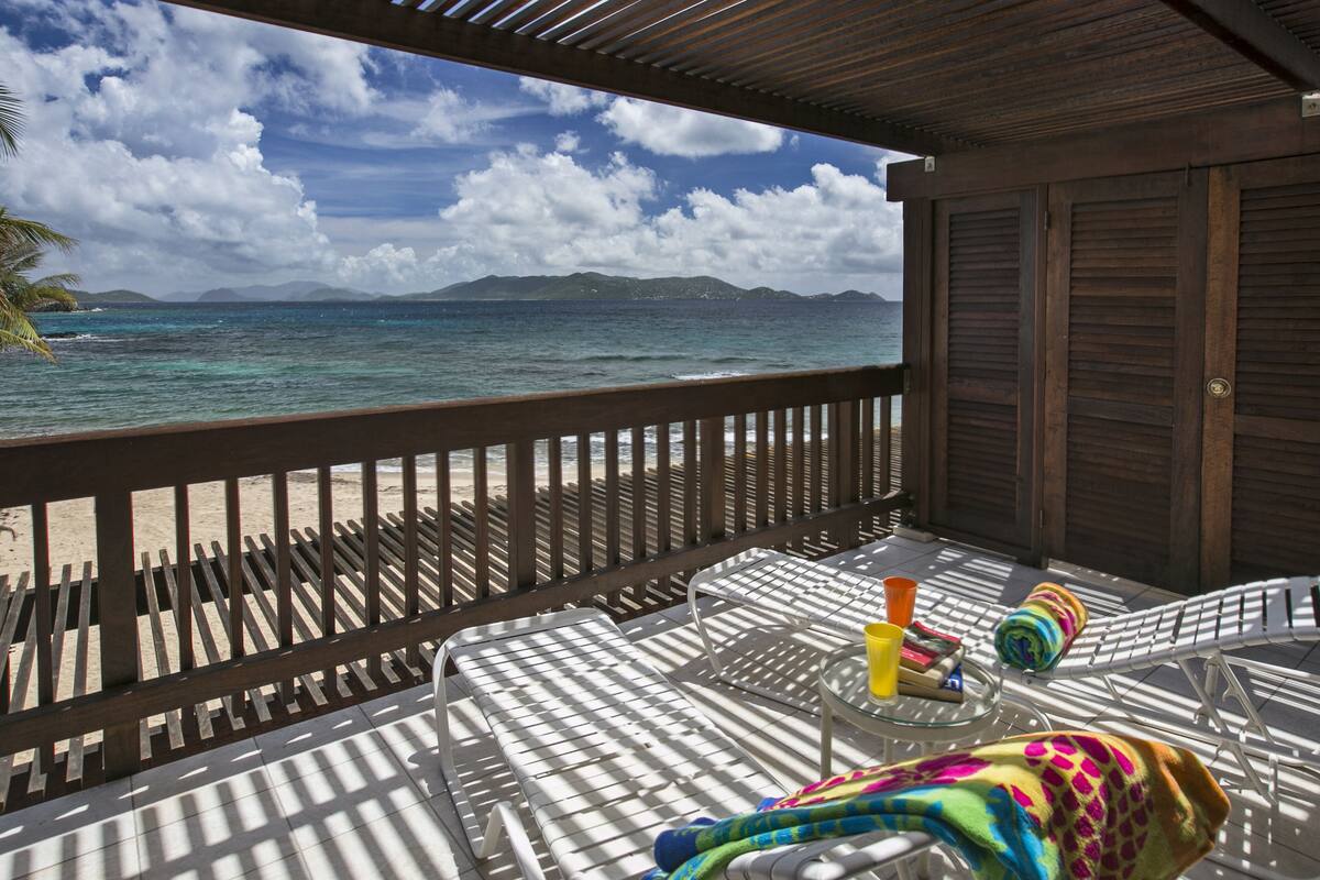 A wooden deck with a view of the ocean and mountains. There are lounge chairs and a small table with a towel and a bottle.