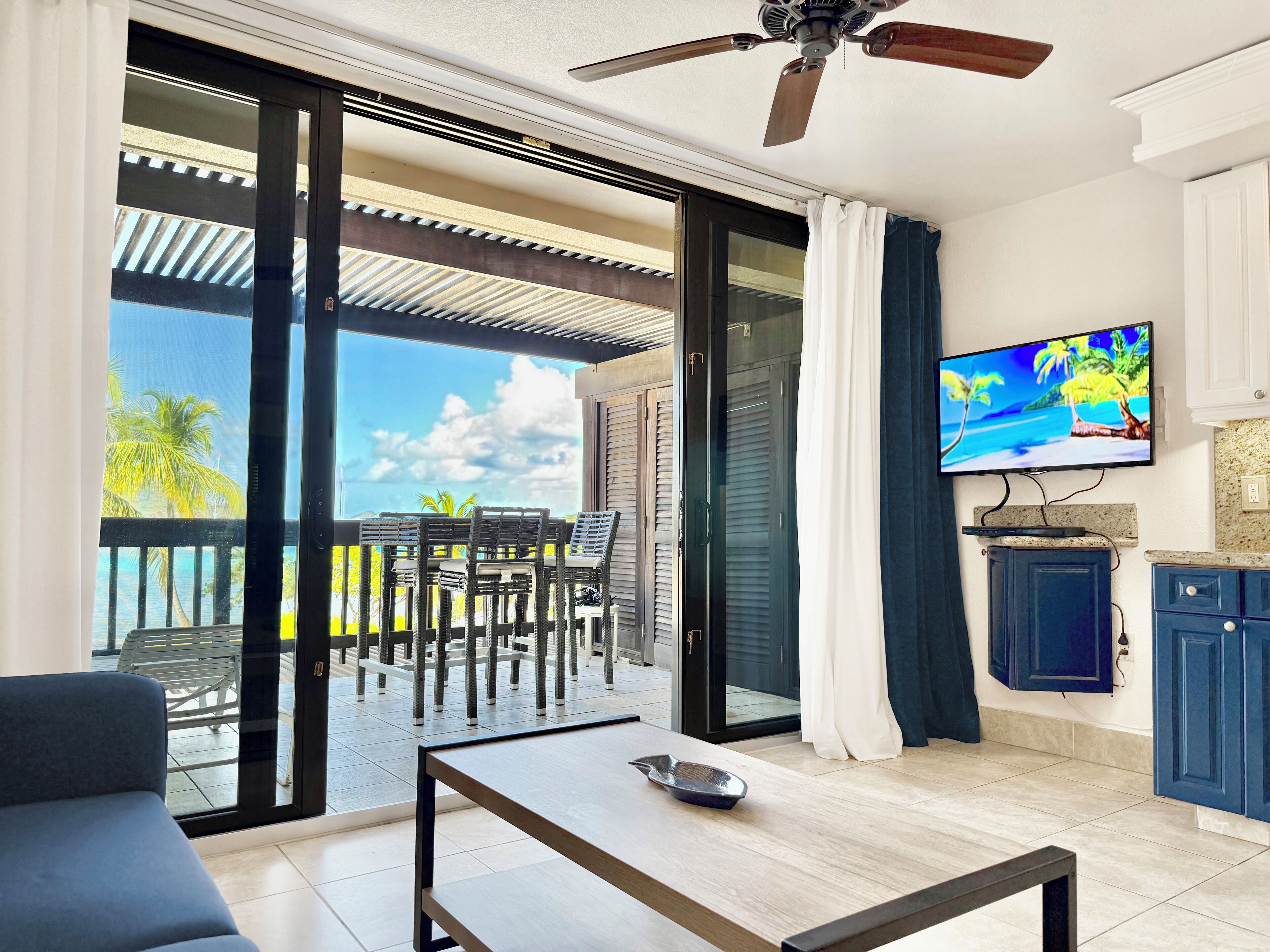 Een moderne woonkamer met een grote schuifpui naar een balkon, een plafondventilator, een flatscreen-televisie en een keuken met blauwe kastjes.