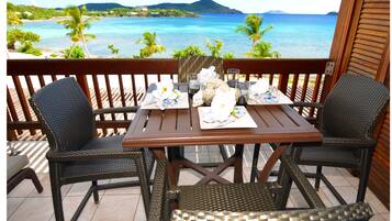 Une table en bois, accompagnée de deux chaises en osier, est placée sur un balcon qui donne sur une plage bordée de palmiers et où l’eau est d’un bleu cristallin.