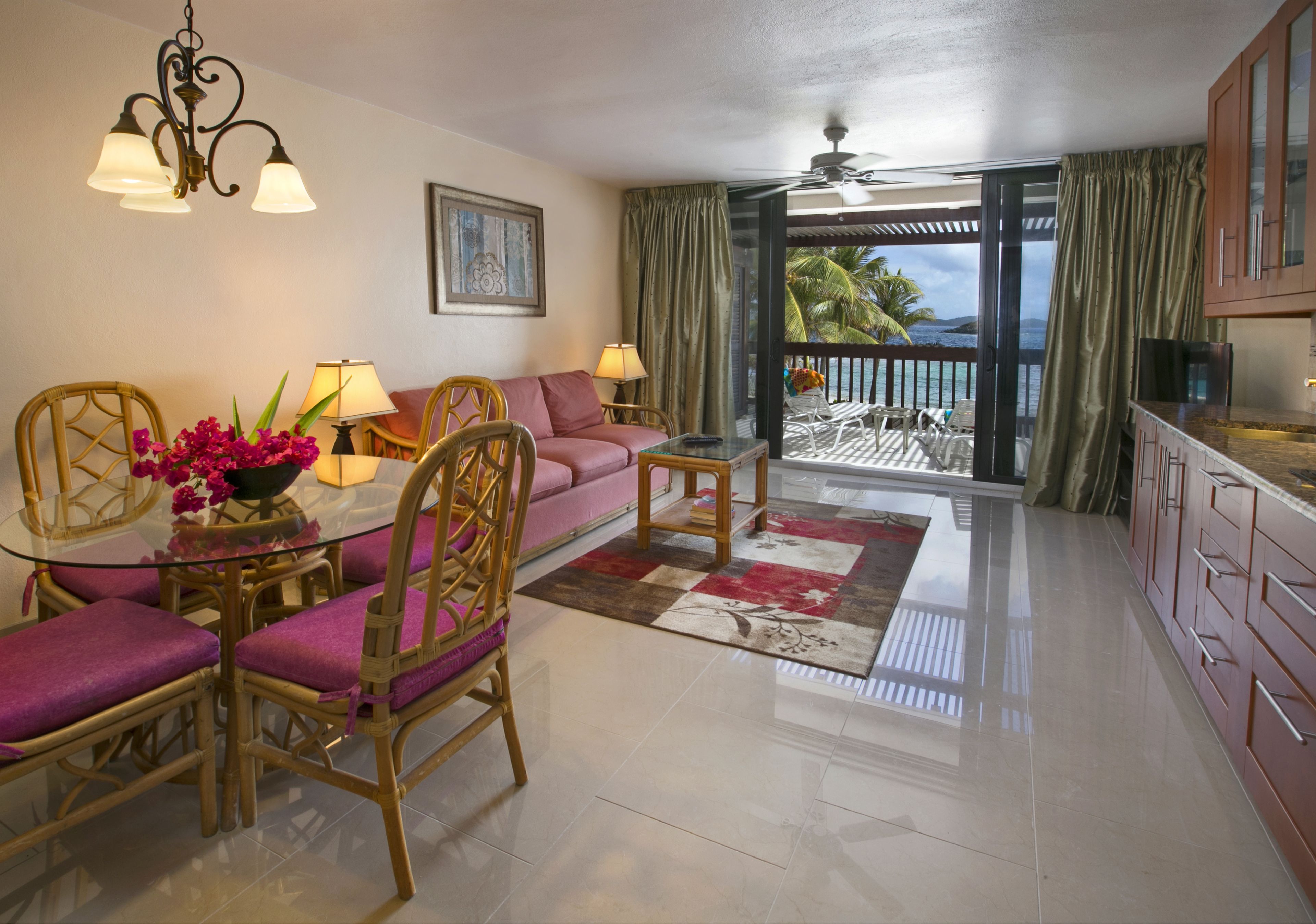 Un coin repas avec une table en verre, des chaises en osier, un canapé rose et une vue sur l’océan, visible à travers une porte coulissante.