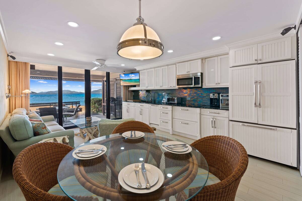 A modern kitchen with white cabinets, a glass dining table, wicker chairs, and a view of the ocean.