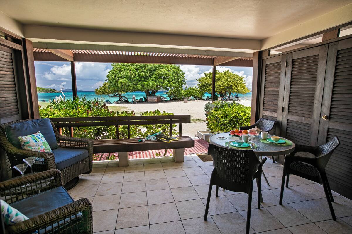 A covered outdoor seating area with wicker furniture, a table with a green placemat, and a view of the beach and ocean.