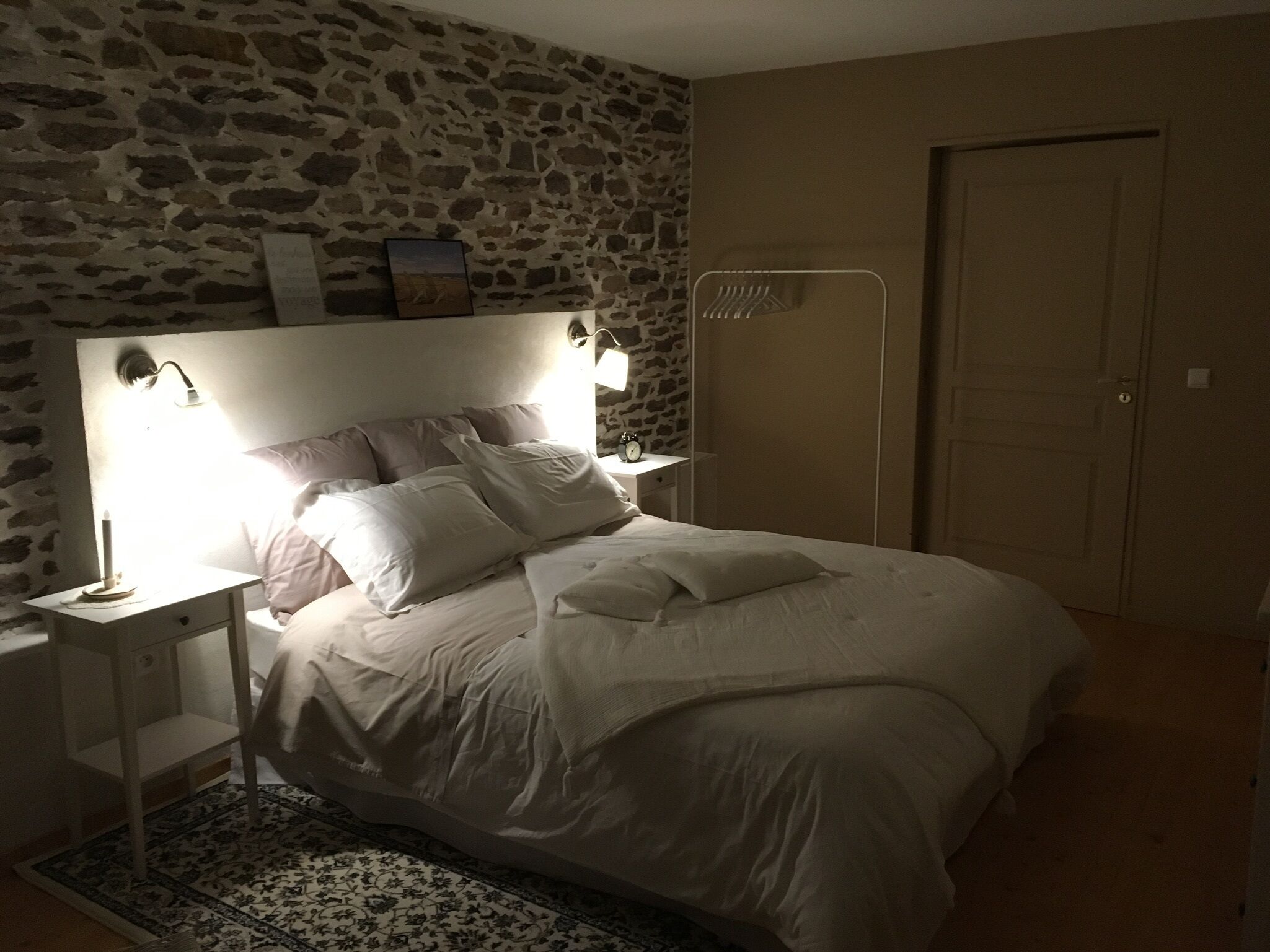 Un dormitorio con pared de piedra, una cama con ropa blanca, una mesita de noche con lámpara y una puerta.