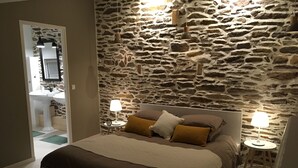 Een slaapkamer met een stenen muur, een bed met bruin beddengoed, twee nachtkastjes met lampen, en een spiegel boven de wastafel in de aangrenzende badkamer.