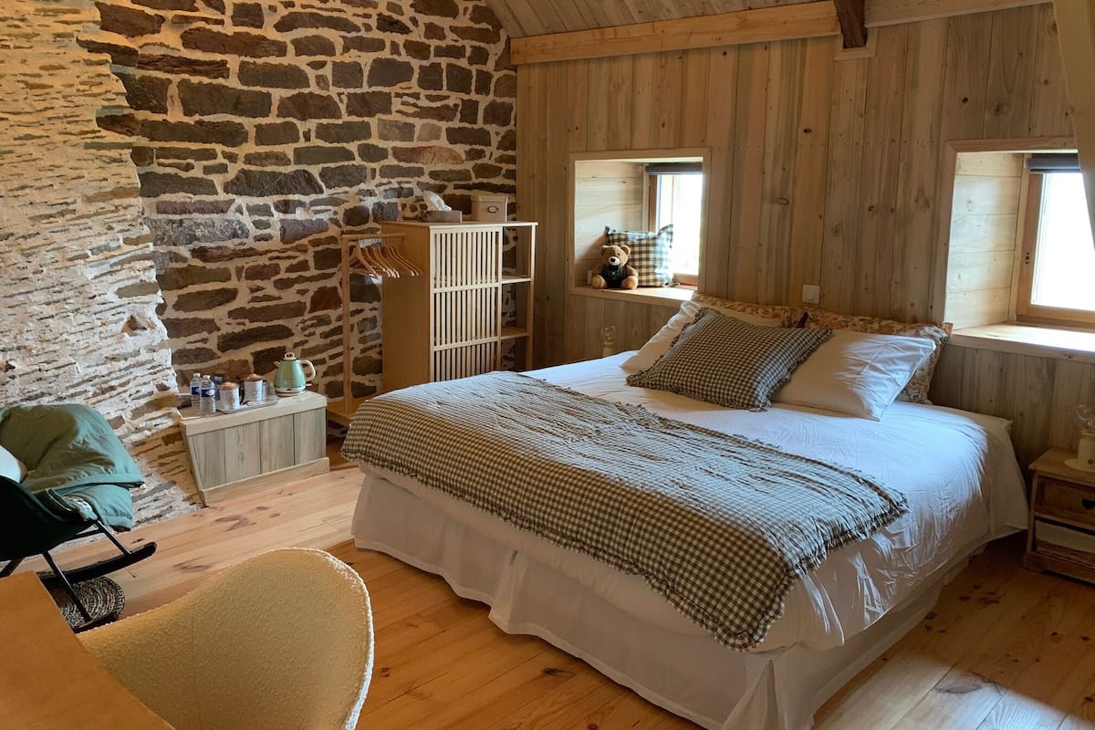 A rustic bedroom with a stone wall, wooden ceiling, a bed with checkered bedding, a bedside table, a chair, and a window with a view.