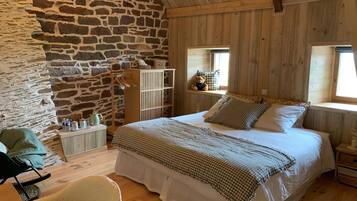 A rustic bedroom with a stone wall, wooden ceiling, a bed with checkered bedding, a bedside table, a chair, and a window with a view.