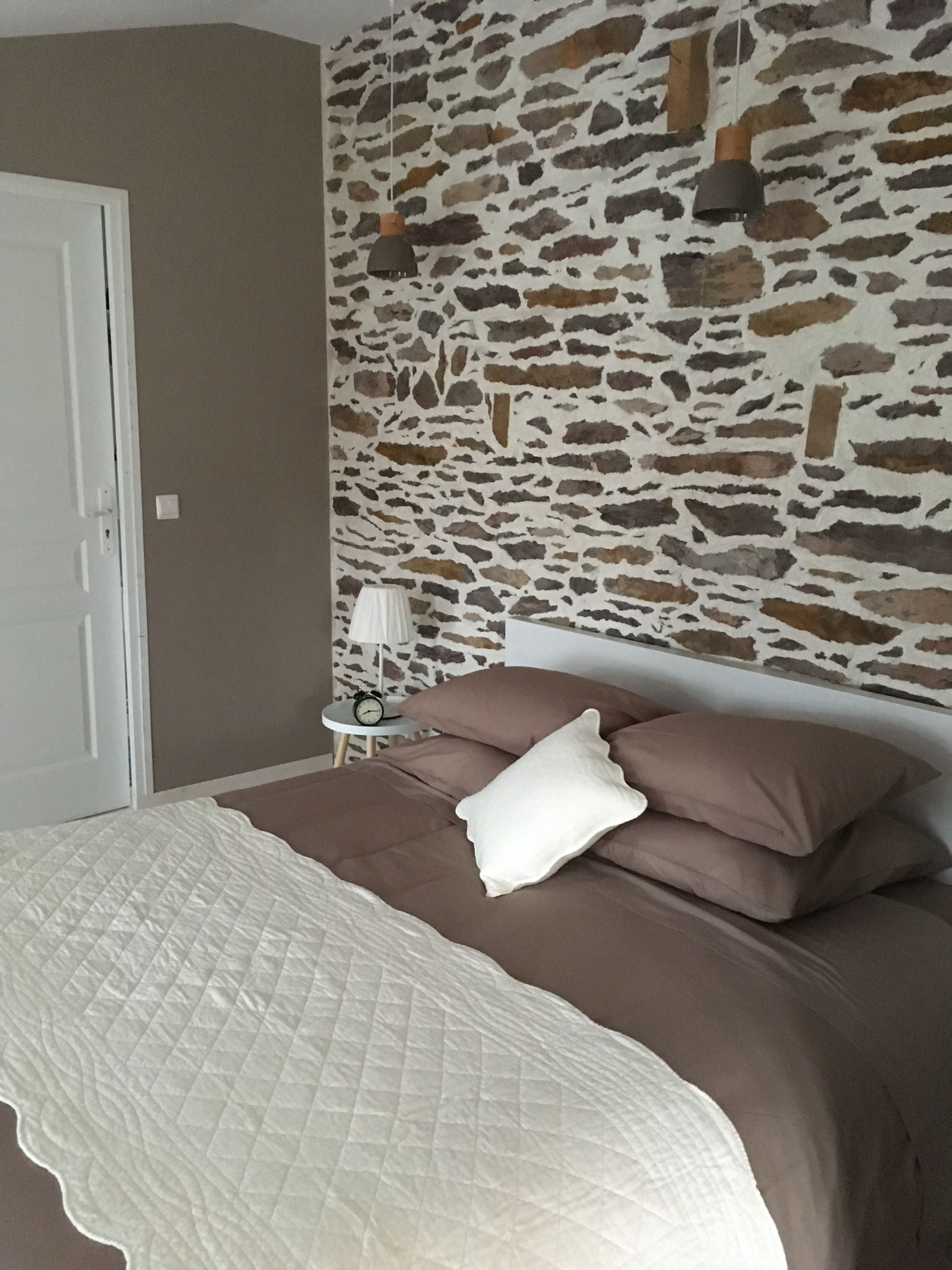 A bedroom with a stone wall, a bed with brown bedding, a white quilted bedspread, and two wall-mounted lamps.