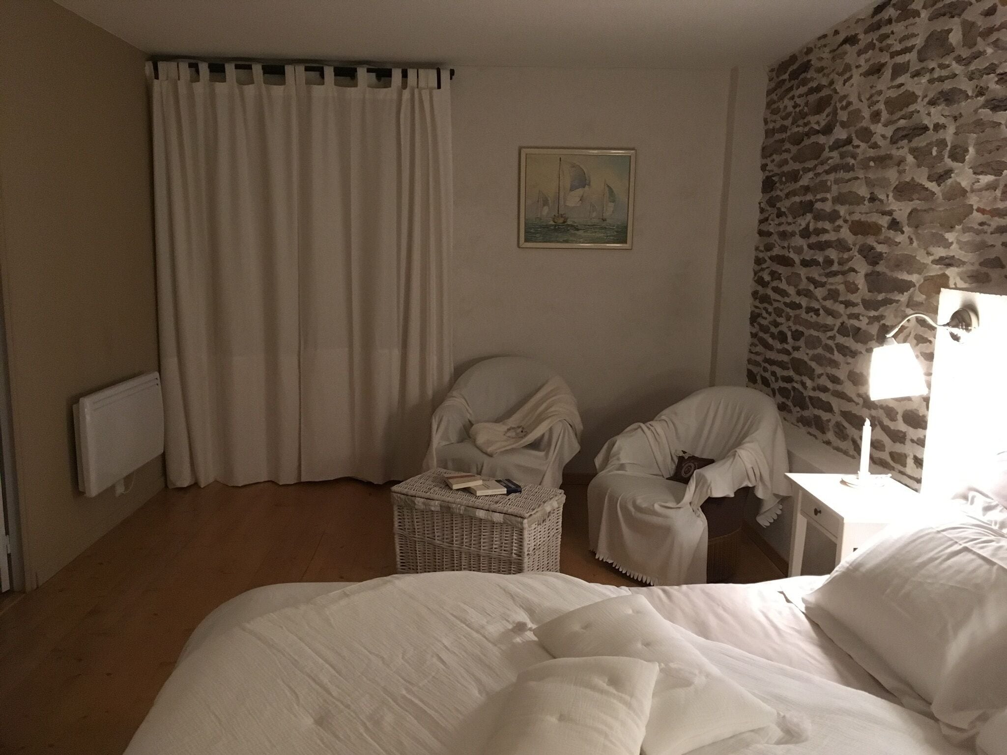 Una habitación de hotel con una cama grande, dos sillones blancos, una cesta de mimbre, un cuadro de veleros, una pared de piedra y una mesita de noche con una lámpara.