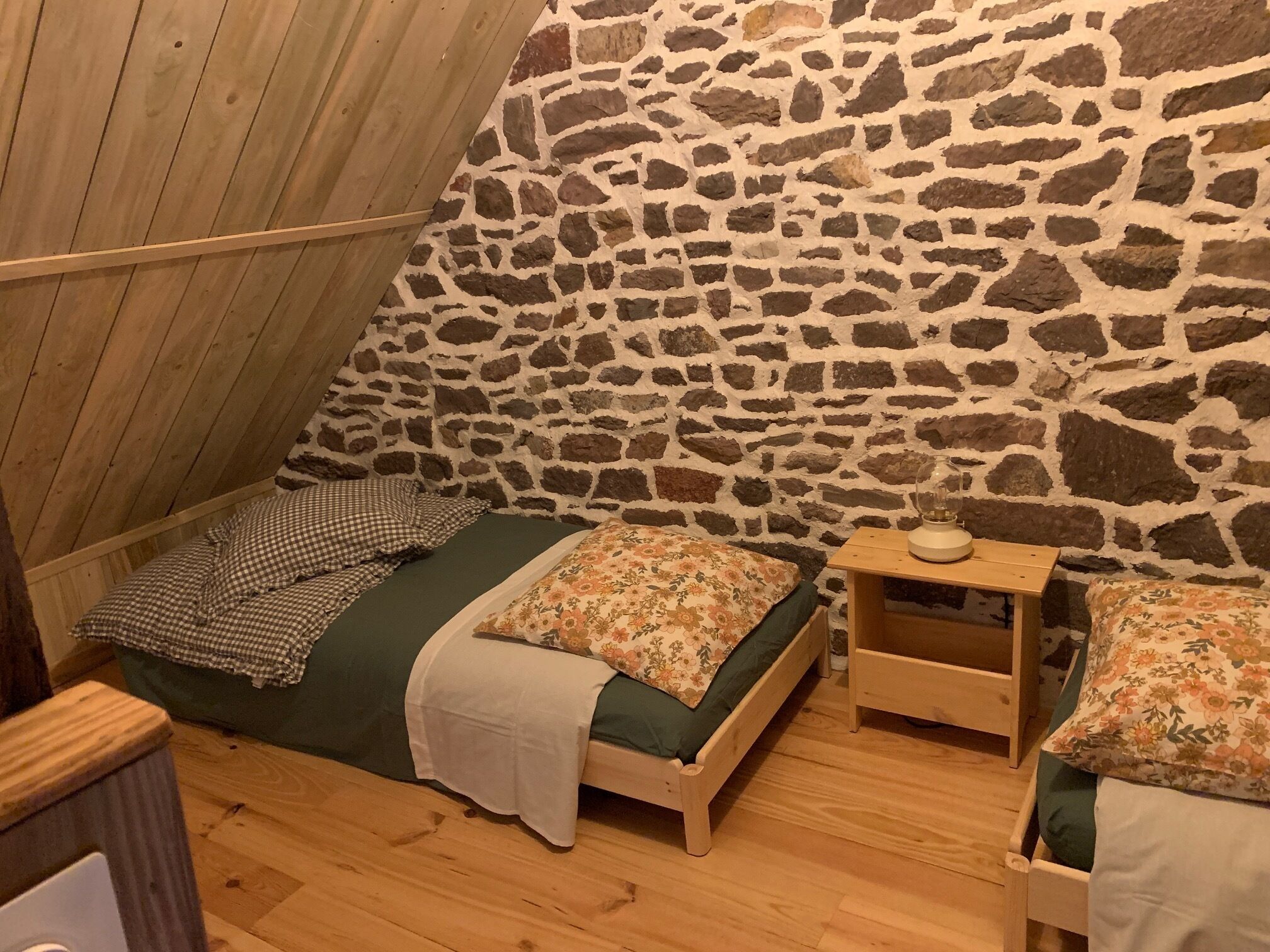 A small, cozy room with a sloped ceiling, stone wall, two wooden beds with floral bedding, and a small wooden nightstand with a lamp.