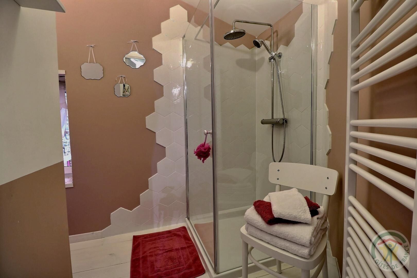 Une douche avec une porte en verre, une chaise blanche avec des serviettes, et une serviette rouge sur le sol.