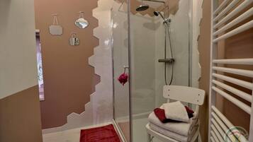Une douche avec une porte en verre, une chaise blanche avec des serviettes, et une serviette rouge sur le sol.