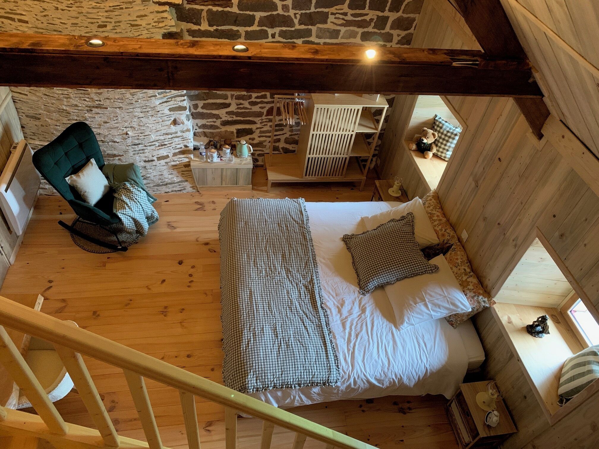 Een slaapkamer in loft-stijl met een hoge plafond, zichtbare houten balken, een stenen muur, een bed met geruit beddengoed, een schommelstoel en een tafeltje met een theepot.