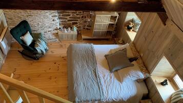 Een slaapkamer in loft-stijl met een hoge plafond, zichtbare houten balken, een stenen muur, een bed met geruit beddengoed, een schommelstoel en een tafeltje met een theepot.