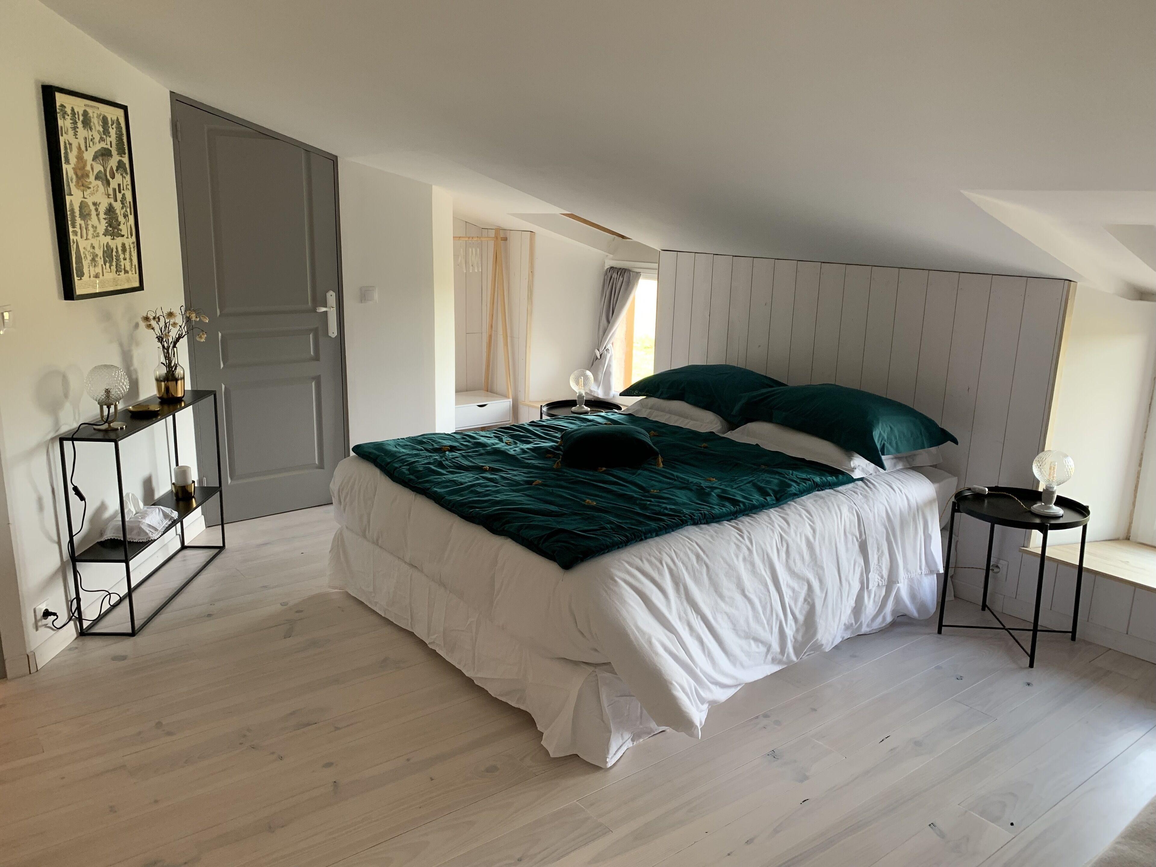 A bedroom with a bed, bedside tables, a console table, a framed botanical print, and a window with curtains.