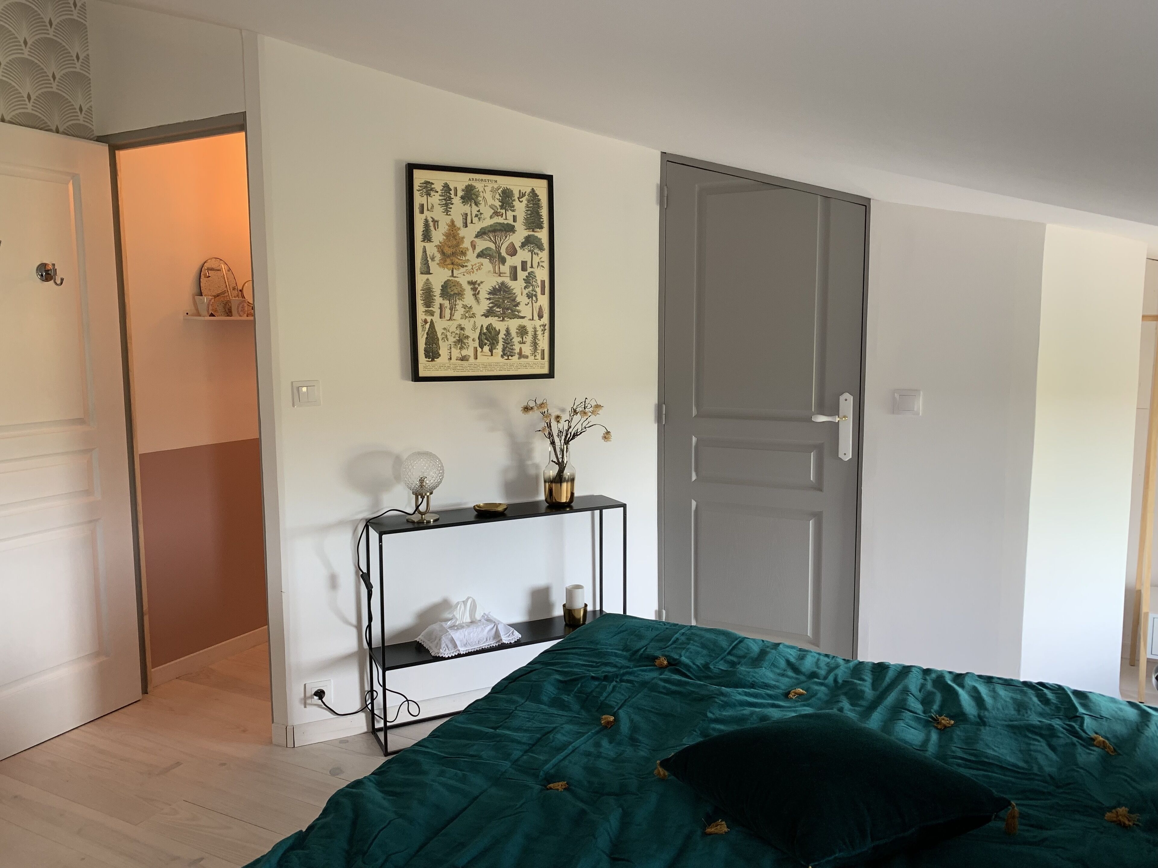 Quarto com colcha verde, uma impressão botânica emoldurada na parede, mesa de cabeceira com abajur e uma porta.