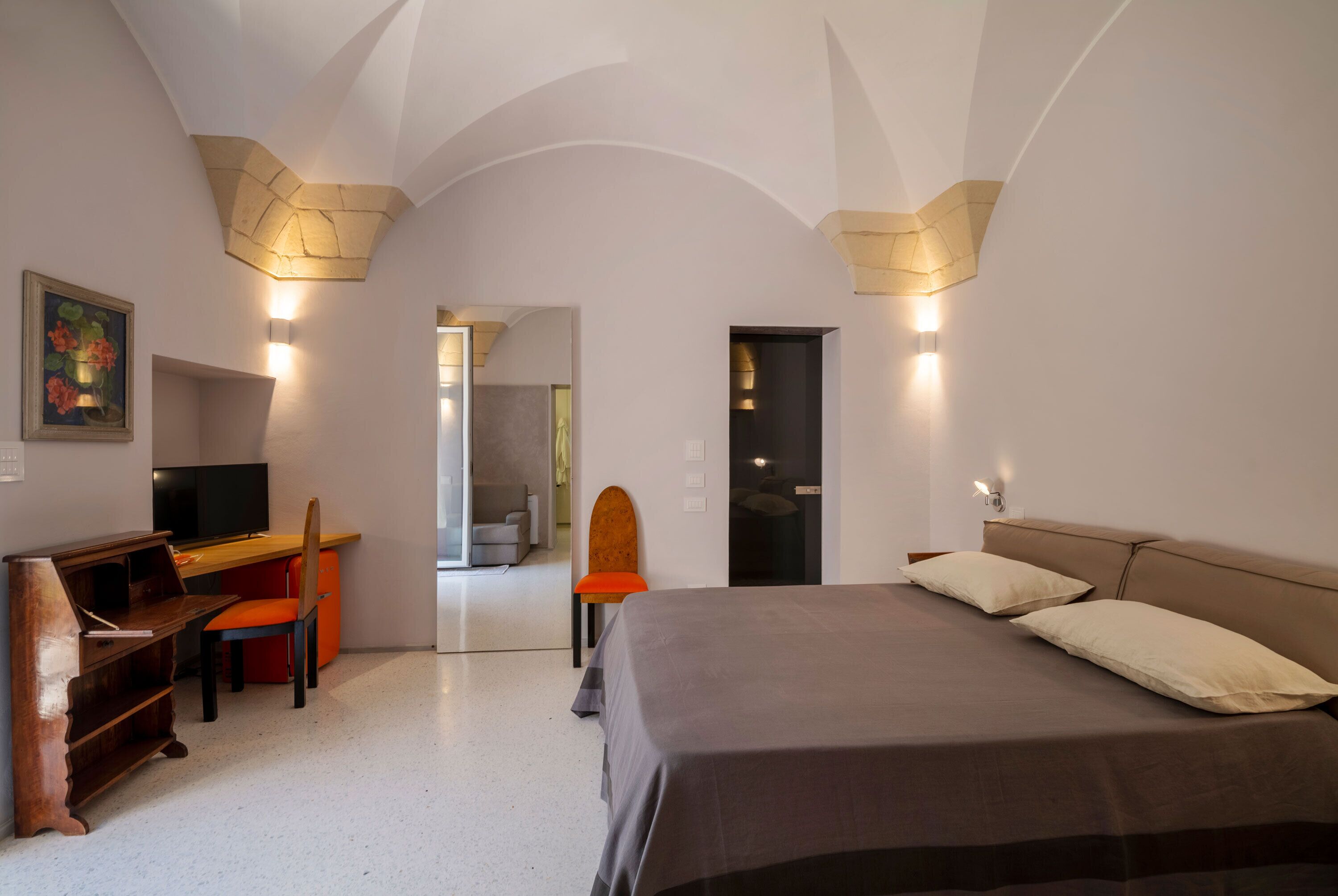 A bedroom with a bed, a desk, and a chair. There is a painting on the wall and a door leading to another room.