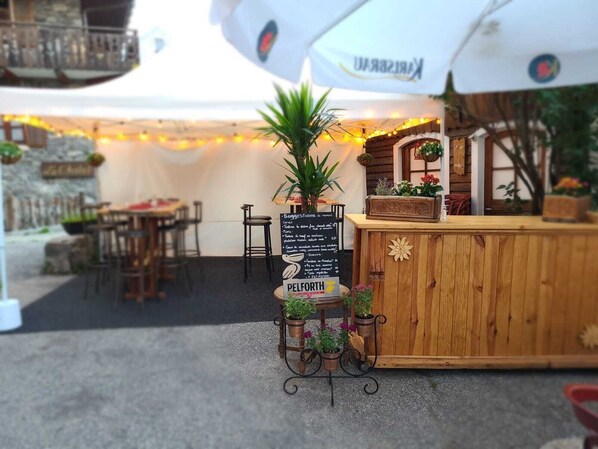 An outdoor seating area with wooden tables and chairs, a signboard with menu items, and a wooden counter with plants.