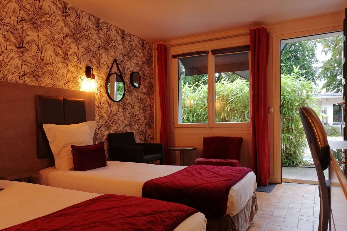Habitación de hotel con dos camas, un ventanal con cortinas, una mesita y una silla.