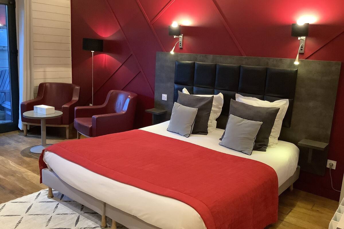 Una habitación de hotel moderna con una cama grande, ropa de cama roja y blanca, una pared de acento roja y dos sillones rojos.