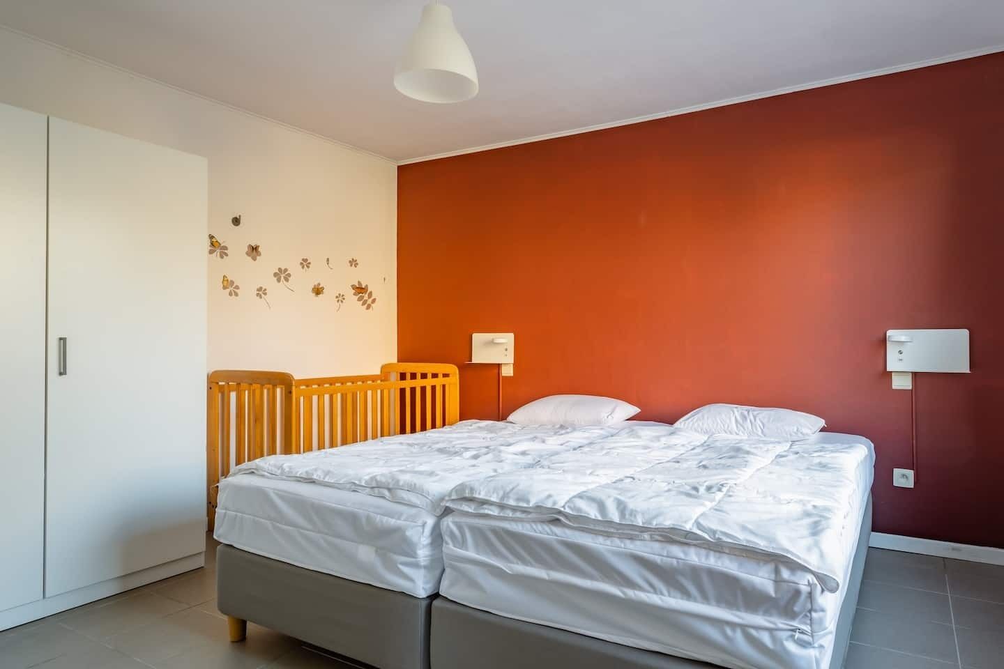Une chambre avec un lit, un berceau en bois, un mur blanc décoré de motifs floraux, un mur d’accent rouge et un luminaire blanc au plafond.