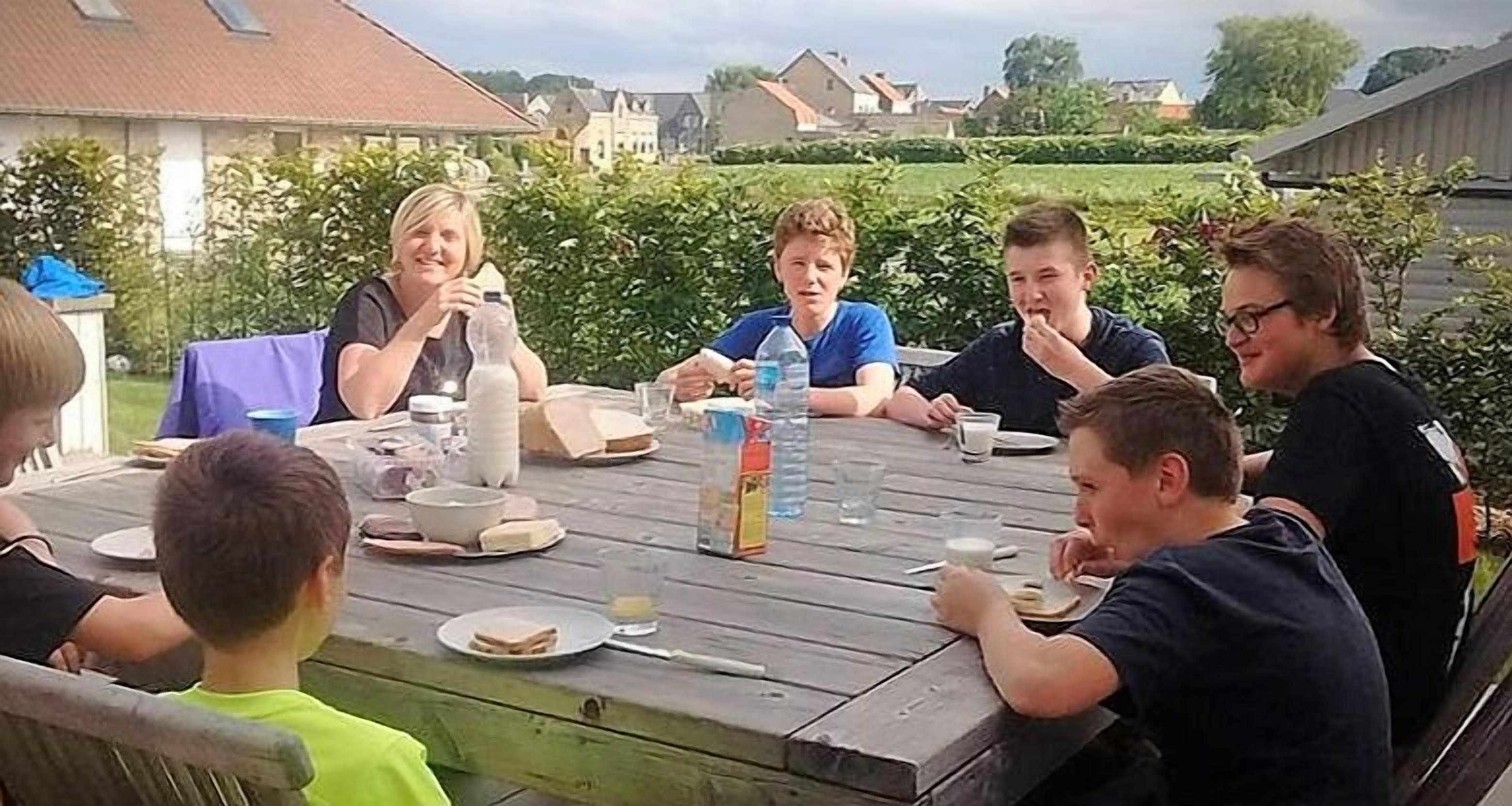A group of people sitting around a wooden table outdoors with a house in the background.