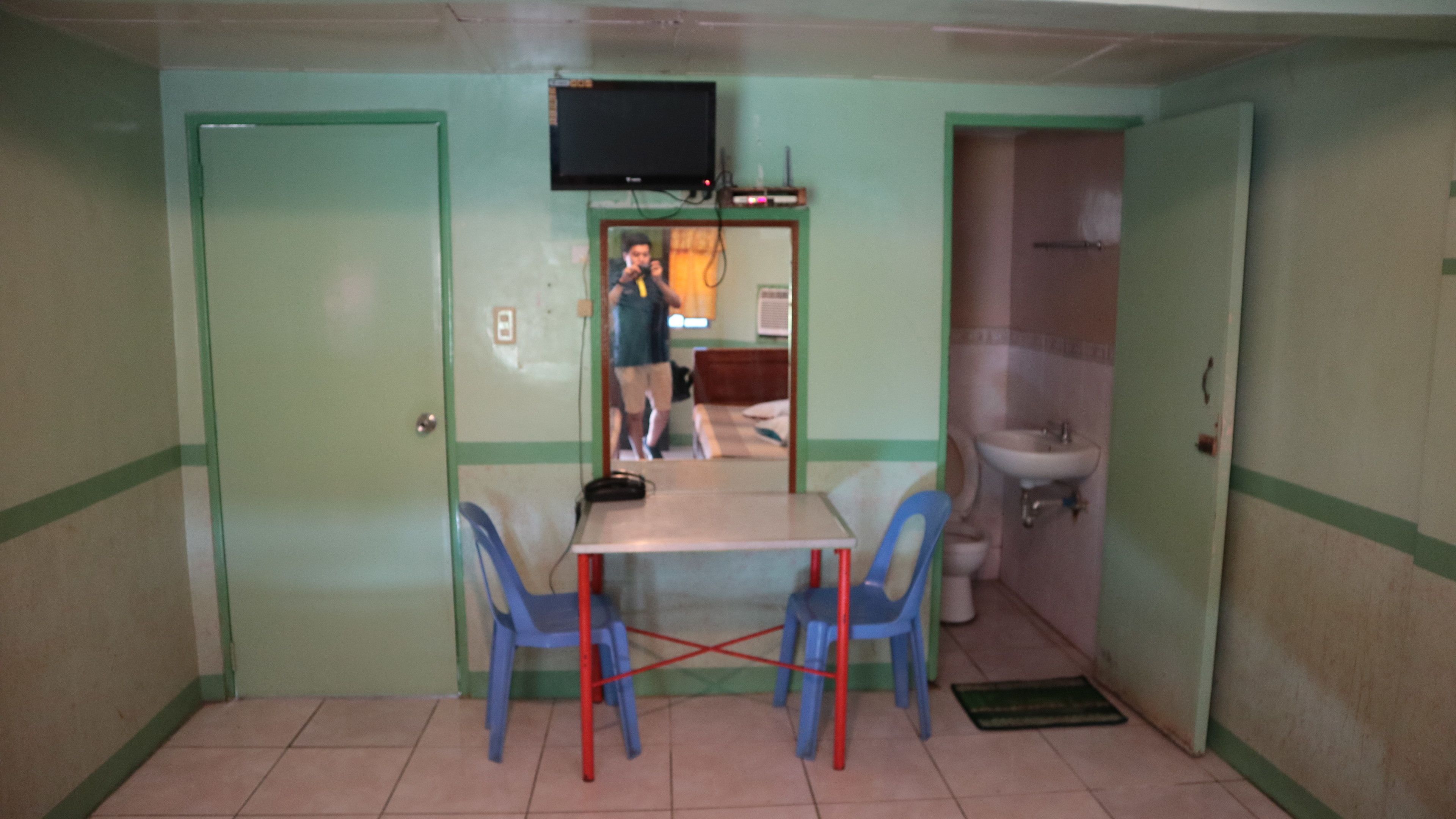 A small room with a table and chairs, a television mounted on the wall, and a bathroom with a sink and toilet.