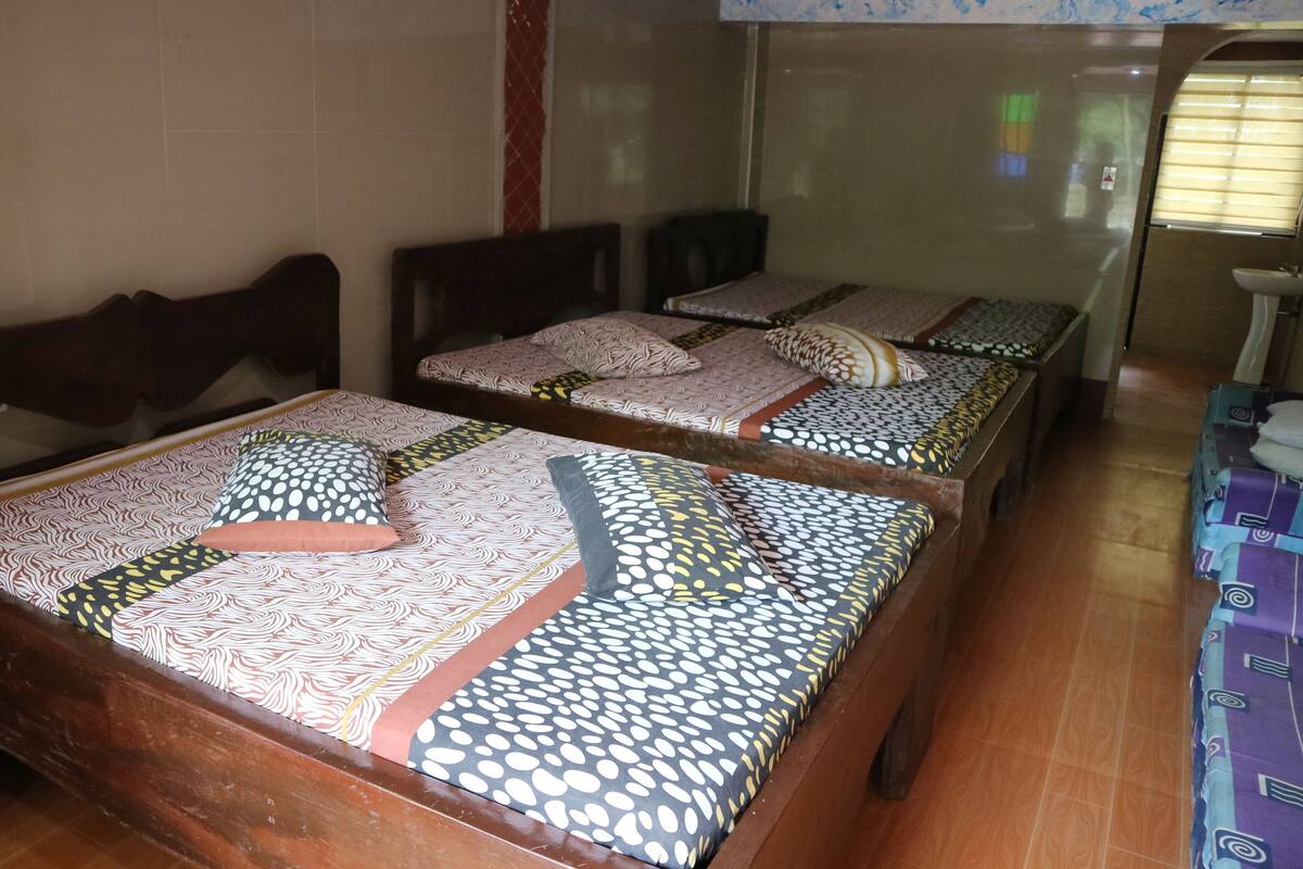 A room with three wooden beds, each with patterned bedding and pillows. The room has a tiled floor and a window with blinds.