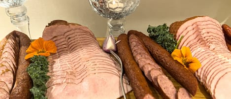 Una mesa de madera con jamón en rodajas, salchichas y flores decorativas.