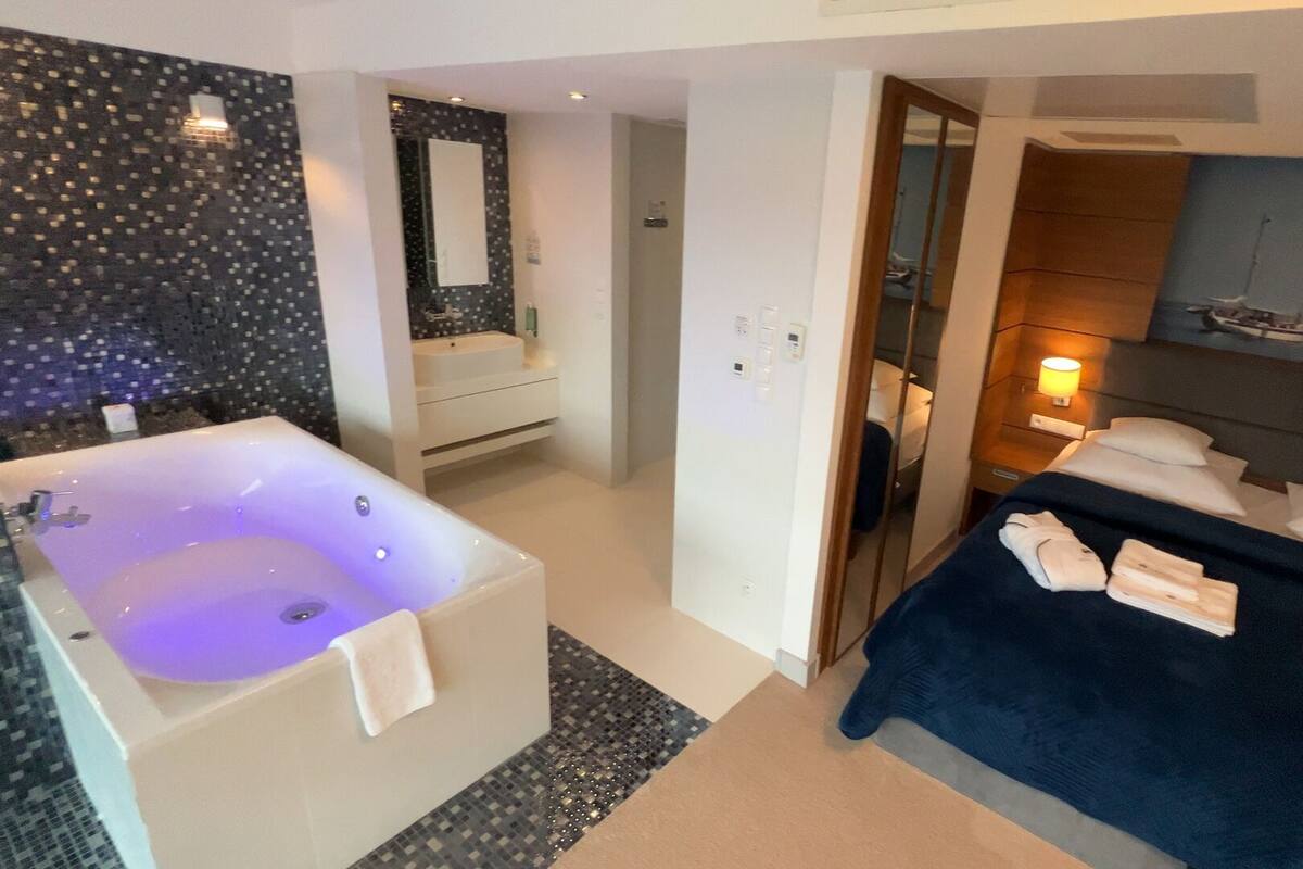 Ein modernes Hotelzimmer mit einer großen Badewanne, einer separaten Duschkabine, einem Bett mit Handtüchern und Meerblick durch das Fenster.