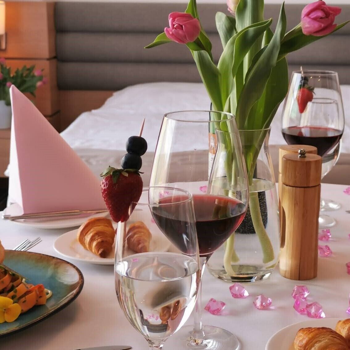 A dining table with wine glasses, a croissant, a strawberry on a skewer, and a vase with pink flowers.