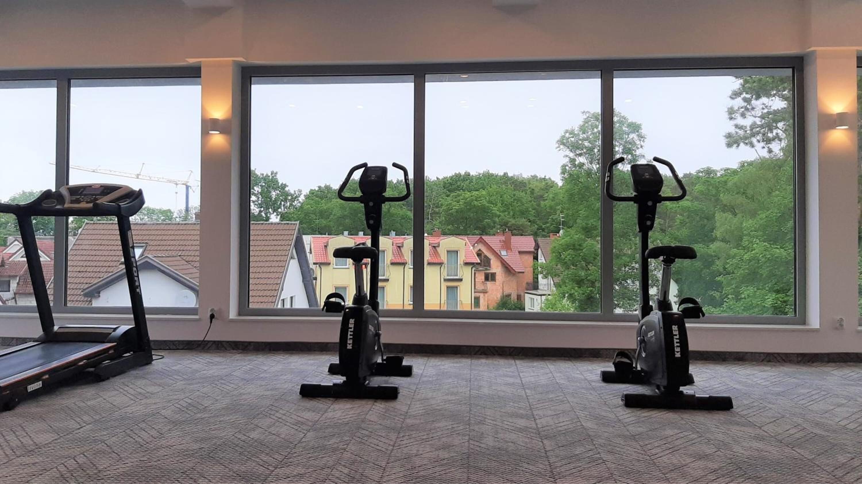 A gym with two stationary bikes and a treadmill, large windows with a view of houses and trees.